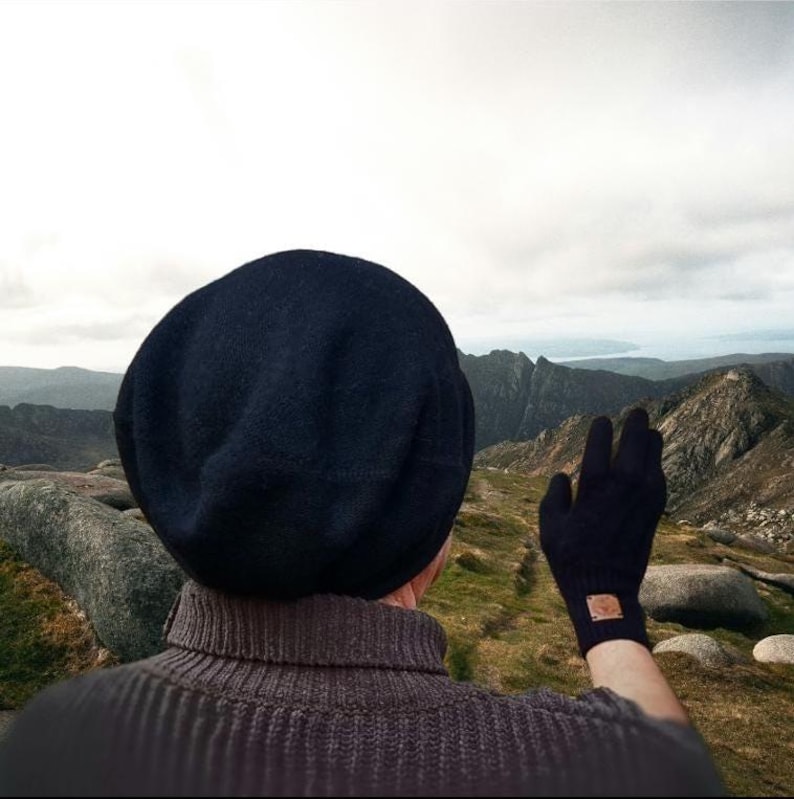 May include: A dark navy blue beanie hat, a dark navy blue glove with a small brown square patch on the wrist, and a dark grey ribbed turtleneck sweater. The items are worn by an individual facing a mountainous landscape under a cloudy sky.