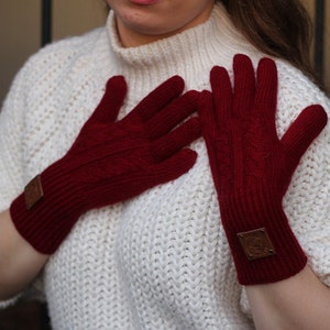 Peut inclure: Une paire de gants en tricot rouge avec un motif en câble et un patch en cuir brun sur chaque gant.