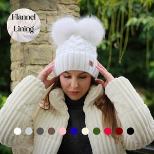 Può includere: Un berretto bianco a trecce con due grandi e soffici pompon. Il cappello ha un'etichetta in pelle marrone ed è foderato in flanella. L'immagine mostra anche una giacca bianca e una tavolozza di colori dei cappelli disponibili.