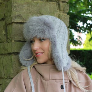 Puede incluir: Un gorro de invierno gris con orejeras y ribete de piel sintética. El gorro está hecho de un material de punto. Las orejeras están diseñadas para mantener las orejas calientes en climas fríos. El gorro tiene una textura suave y esponjosa.