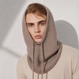 May include: A light brown, ribbed knit balaclava hood featuring a fitted design that frames the face. The head covering is crafted from a soft, textured material and includes two long, matching knit drawstrings hanging down from the front neckline.