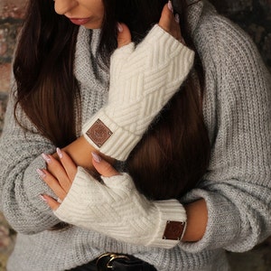 Peut inclure: Une paire de gants sans doigts blanc cassé avec un motif en tricot torsadé texturé et un poignet côtelé. Chaque gant est orné d'un petit écusson carré en similicuir marron gaufré d'un logo circulaire et de clous métalliques aux coins.