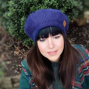May include: A woman wearing a purple beret with a brown leather patch. She has long dark hair and is wearing a green and red plaid shirt.