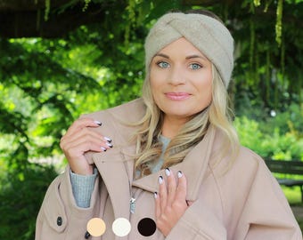May include: A woman wearing a beige wool headband and a matching coat. The headband has a twisted design. The coat has a zipper closure and a large collar. The background is a lush green park.