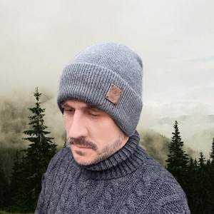 May include: A gray knit beanie with a brown leather patch. The hat is ribbed and has a folded brim. The person in the image is wearing a gray cable knit sweater. The background features a mountain landscape.