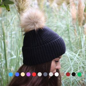Könnte beinhalten: Eine schwarze Strickmütze mit einem großen, flauschigen, hellbraunen Bommel. Die Mütze ist gerippt und scheint aus einem weichen Material zu sein. Der Hintergrund zeigt hohes grünes Gras.