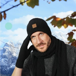May include: A black knitted beanie with a brown leather patch, a black knitted scarf, and black knitted gloves. The person is wearing a grey sweater and is standing in front of a snowy mountain range.