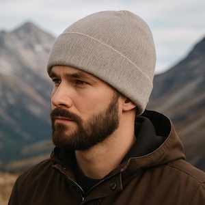 May include: A light grey beanie hat with a folded brim. The hat is made of a knitted material. The person in the image is wearing a brown jacket. The background features mountains and a cloudy sky.
