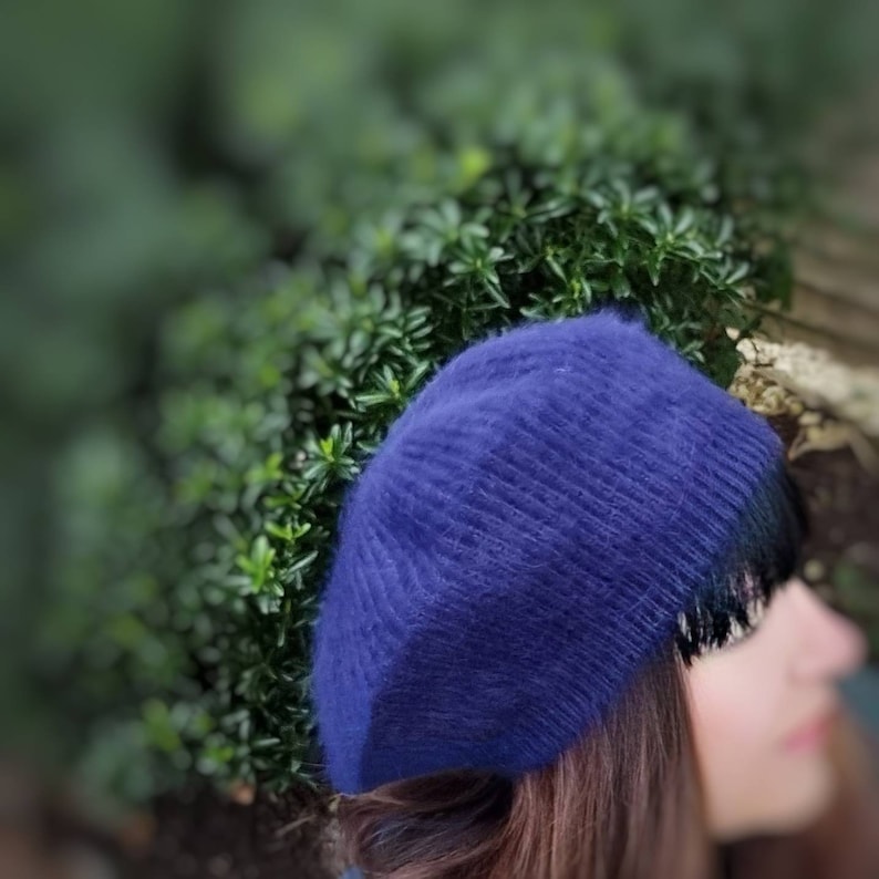 May include: A blue knitted beret with a fringe detail. The beret is made of a soft, fuzzy material.