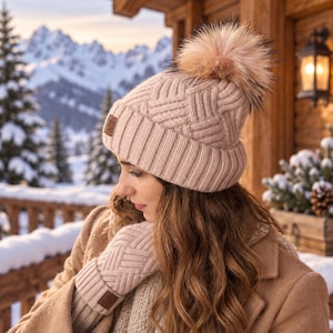 May include: A light pink knit beanie with a matching pair of knit mittens. The beanie features a textured basketweave pattern, a wide ribbed cuff, and a large, fluffy faux fur pom-pom on top. Both items include a small, square brown leather tag.