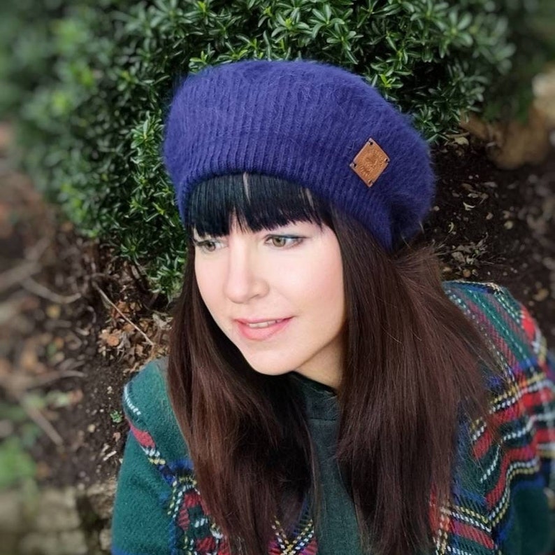May include: A woman wearing a dark blue fuzzy beret with a brown leather patch. She is wearing a green, red, and black plaid shirt.