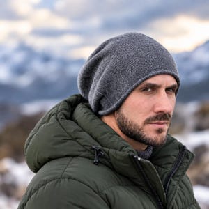 May include: A charcoal grey knit beanie hat with a soft, textured surface and a slightly slouchy fit. The beanie is constructed from a fine-gauge knit fabric, providing a simple, rounded silhouette that covers the head comfortably.