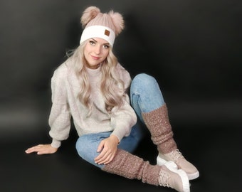 May include: A woman wearing a white and pink knitted beanie with ear flaps and a pair of pink and white leg warmers. She is wearing a light pink sweater and blue jeans.
