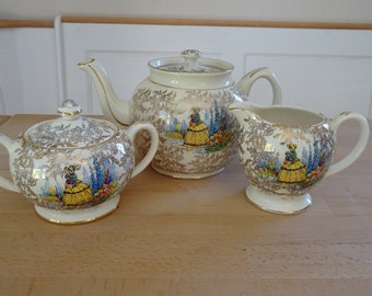 May include: A vintage tea set with a cream-colored base and gold accents. The teapot, sugar bowl, and creamer feature a floral pattern with a lady in a yellow dress.