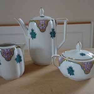 Op de afbeelding: Een wit porseleinen theeservies met een gouden rand en groene en paarse bloemenmotieven. De set bevat een theepot, een roomkan en een suikerpot.