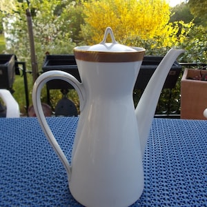 Może przedstawiać: Biały ceramiczny dzbanek do kawy z złotą obwódką wokół pokrywki i złotą wstążką wokół podstawy. Dzbanek ma długi dzióbek i uchwyt.