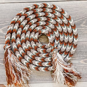 May include: A coiled rope with a braided pattern in shades of brown, white, and green. The rope has decorative tassels at each end, adding a rustic touch. The rope is designed for equestrian use.