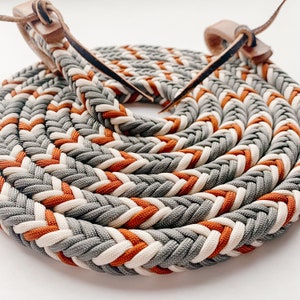 May include: A braided rope with a grey, white, and orange color pattern. The rope is coiled and has leather ends.
