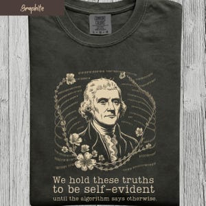 May include: A graphite gray t-shirt featuring a cream-colored graphic of Thomas Jefferson surrounded by floral vines and binary code. The text reads: We hold these truths to be self-evident until the algorithm says otherwise.