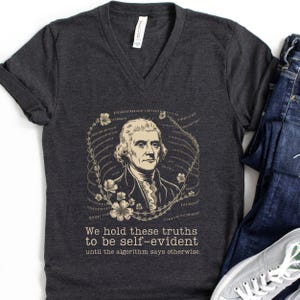 May include: A dark heather gray V-neck t-shirt featuring a graphic of Thomas Jefferson surrounded by binary code and flowers. The text reads: We hold these truths to be self-evident until the algorithm says otherwise. The shirt is paired with blue jeans.