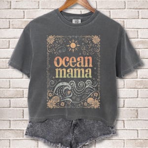 May include: Dark gray t-shirt with the text "Ocean Mama" in a colorful, vintage-style font. The design includes a sun, waves, and floral elements. The shirt is hanging on a wooden hanger.