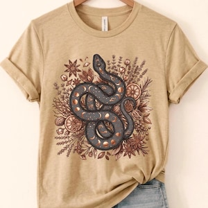 May include: A tan short-sleeve t-shirt featuring a central graphic of a dark grey coiled snake adorned with copper-coloured moon phases and planetary symbols, surrounded by detailed illustrations of dried botanicals, star anise, and citrus slices.
