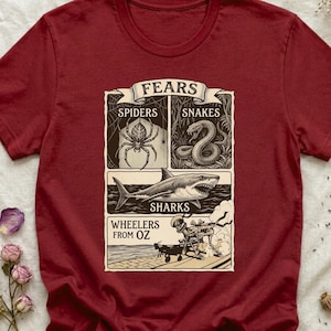May include: Maroon t-shirt featuring a graphic design with the word "FEARS" at the top. The design includes illustrations of a spider, snake, shark, and a scene from "The Wizard of Oz".