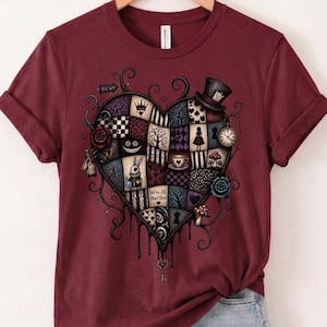 May include: A maroon short-sleeve t-shirt featuring a large heart-shaped graphic composed of various patterned patches. The design includes a rabbit, a top hat, a pocket watch, a teacup, and the text "We're All Mad Here" with black dripping accents.
