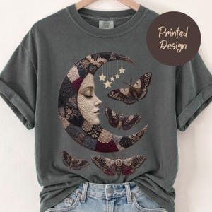 May include: A charcoal gray crew neck t-shirt featuring a graphic of a crescent moon with a human face, decorated with a patchwork pattern in shades of burgundy, black, and cream. Four moths and five hanging stars surround the moon design.