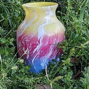 May include: A colorful glass vase with a unique design. The vase transitions from yellow at the top to pink, white, and blue at the bottom. The colors blend with a marbled effect. The vase is surrounded by green grass.