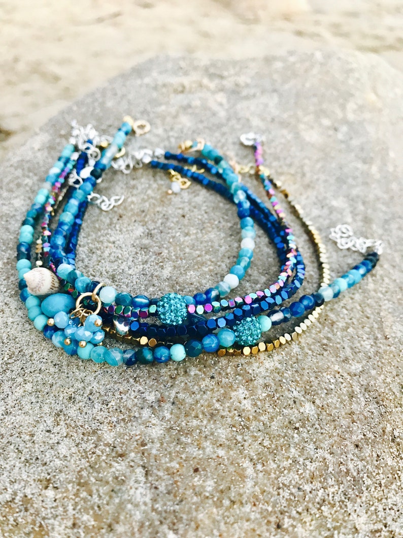 K&ouml;nnte beinhalten: Mehrere geschichtete Perlenarmb&auml;nder in verschiedenen Blau- und Goldt&ouml;nen. Die Armb&auml;nder weisen eine Mischung aus Perlengr&ouml;&szlig;en und -strukturen auf, darunter schillernde und strukturierte Perlen sowie ein kleiner Muschelanh&auml;nger. Die Armb&auml;nder werden auf einer strukturierten, hellfarbigen Oberfl&auml;che pr&auml;sentiert.