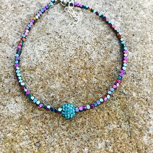 K&ouml;nnte beinhalten: Ein schillerndes Perlenarmband mit einer zentralen, t&uuml;rkisfarbenen, funkelnden Perle. Das Armband hat kleine, quadratische Perlen in einer Reihe von Farben, darunter Lila, Gr&uuml;n und Blau. Der Verschluss ist silberfarben.