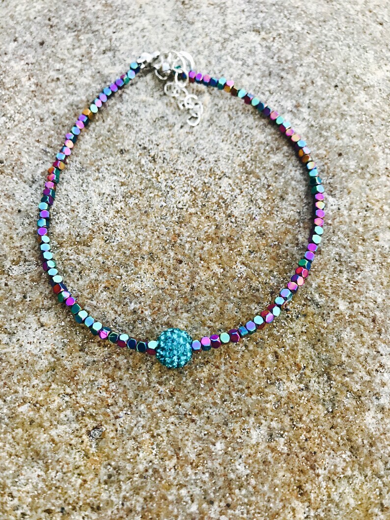K&ouml;nnte beinhalten: Ein schillerndes Perlenarmband mit einer zentralen, t&uuml;rkisfarbenen, funkelnden Perle. Das Armband hat kleine, quadratische Perlen in einer Reihe von Farben, darunter Lila, Gr&uuml;n und Blau. Der Verschluss ist silberfarben.