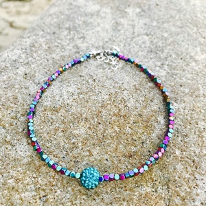 K&ouml;nnte beinhalten: Ein zartes, schillerndes Perlenarmband mit einer zentralen t&uuml;rkisfarbenen, strukturierten Perle. Die Perlen des Armbands zeigen ein Farbspektrum, darunter Lila, Gr&uuml;n und Blau. Der Verschluss ist silberfarben. Das Armband liegt auf einer strukturierten, hellen Oberfl&auml;che.