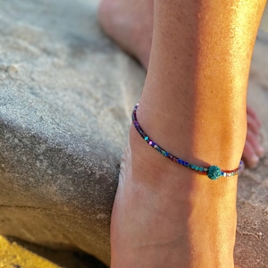 K&ouml;nnte beinhalten: Fu&szlig;kettchen mit kleinen, farbenfrohen Perlen in Blau-, Lila- und Gr&uuml;nt&ouml;nen. Eine gr&ouml;&szlig;ere, t&uuml;rkis farbene Perle befindet sich in der Mitte des Armbands. Der Schmuck wird am Kn&ouml;chel getragen.
