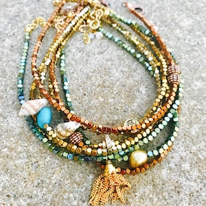 K&ouml;nnte beinhalten: Eine Sammlung von Armb&auml;ndern mit Perlen in Gold-, Kupfer- und Gr&uuml;nt&ouml;nen, verziert mit Muscheln, einer t&uuml;rkisfarbenen Perle und einer goldenen Quaste. Die Armb&auml;nder sind geschichtet und erzeugen eine strukturierte und farbenfrohe Darstellung.