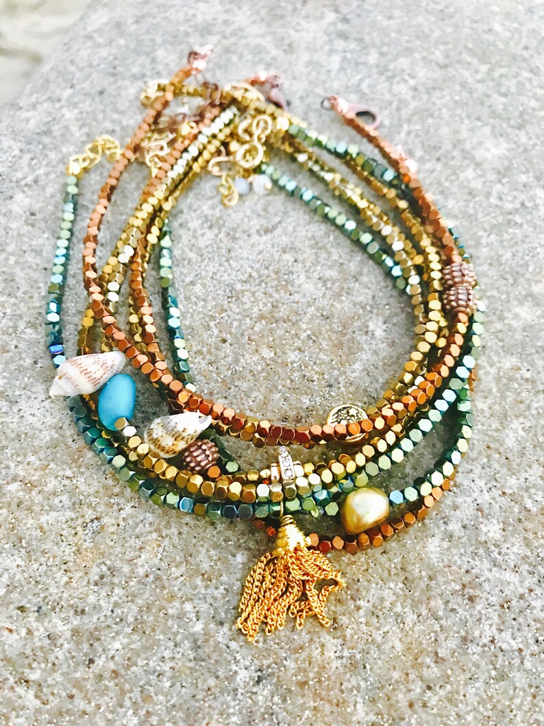 K&ouml;nnte beinhalten: Eine Sammlung von Armb&auml;ndern mit Perlen in Gold-, Kupfer- und Gr&uuml;nt&ouml;nen, verziert mit Muscheln, einer t&uuml;rkisfarbenen Perle und einer goldenen Quaste. Die Armb&auml;nder sind geschichtet und erzeugen eine strukturierte und farbenfrohe Darstellung.