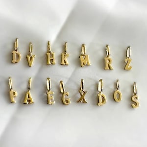 Può includere: Ciondoli con alfabeto color oro, ciascuno con un piccolo anello per il fissaggio. Sono visualizzate le lettere D, V, H, K, M, R, Z, P, A, E, G, X, D, O e S. I ciondoli sono disposti su una superficie bianca.