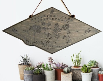 May include: A wooden Ouija board with a gray stain and black lettering. The board features a diamond shape with the letters of the alphabet, numbers 1-9 and 0, the words "yes", "no", and "farewell", and a snake design. The board is hanging from a brown cord.