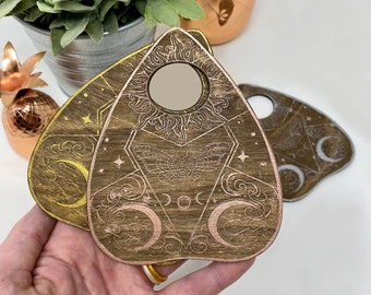 May include: A wooden Ouija board with a gold and rose gold finish. The board features a crescent moon, stars, and a moth design. The board has a small mirror in the centre.