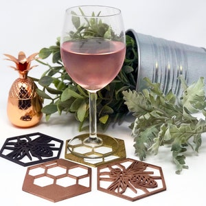 May include: Four hexagonal coasters with a bee design. Two coasters are brown, one is black, and one is gold. A glass of pink wine sits on the gold coaster. A small copper pineapple sits on the table.