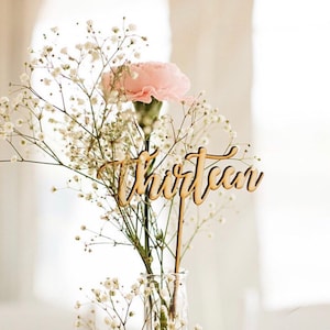 May include: Wooden table number with the word "thirteen" written in cursive. The number is placed in a vase with white baby's breath and a pink carnation.
