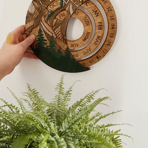Peut inclure: Un calendrier en bois avec un motif de montagne et de for&ecirc;t. Le calendrier a la forme d'un croissant de lune et poss&egrave;de un cadran circulaire avec les jours de la semaine et les mois de l'ann&eacute;e. Le calendrier est en bois et a une finition brun fonc&eacute;.