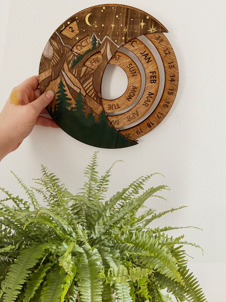Peut inclure: Un calendrier en bois avec un motif de montagne et de for&ecirc;t. Le calendrier a la forme d'un croissant de lune et poss&egrave;de un cadran circulaire avec les jours de la semaine et les mois de l'ann&eacute;e. Le calendrier est en bois et a une finition brun fonc&eacute;.