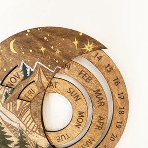 Peut inclure: Calendrier en bois avec une sc&egrave;ne de montagne et un ciel &eacute;toil&eacute;. Le calendrier a un cadran rotatif avec les mois de l'ann&eacute;e et les jours de la semaine. Les num&eacute;ros 14 &agrave; 20 sont visibles sur le cadran.