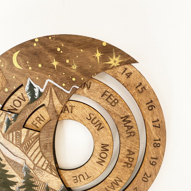 Peut inclure: Calendrier en bois avec une sc&egrave;ne de montagne et un ciel &eacute;toil&eacute;. Le calendrier a un cadran rotatif avec les mois de l'ann&eacute;e et les jours de la semaine. Les num&eacute;ros 14 &agrave; 20 sont visibles sur le cadran.