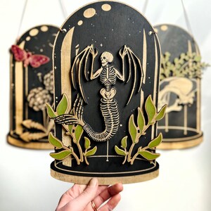 May include: A wooden wall hanging with a black background and a skeletal mermaid inside a glass dome. The mermaid has wings and a tail. The dome is surrounded by green leaves.