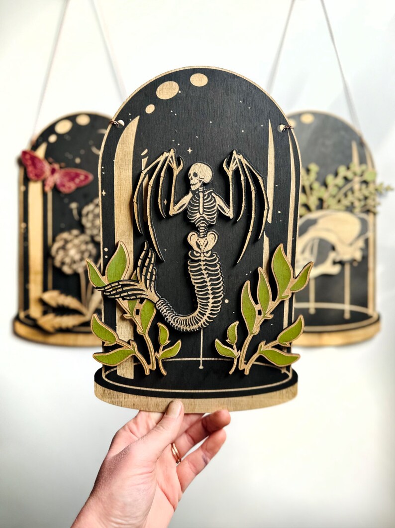 May include: A wooden wall hanging with a black background and a skeletal mermaid inside a glass dome. The mermaid has wings and a tail. The dome is surrounded by green leaves.