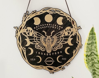 May include: A wooden Ouija board with a black background and gold lettering. The board features a moth design, moon phases, and the letters of the alphabet, numbers 1-9 and 0, and the words "YES", "FAREWELL", and "NO".