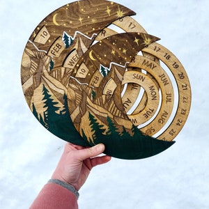 Peut inclure: Calendrier en bois avec un motif de montagne et de for&ecirc;t. Le calendrier a la forme d'un croissant de lune et poss&egrave;de un cadran rotatif pour chaque mois. Le calendrier est en bois et a une palette de couleurs verte et brune.